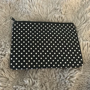 Svart prickig kuvertväska med dragkedja - Svart kuvertväska med vita prickar och praktisk dragkedja upptill. Perfekt storlek för att ha med mobilen, smink eller andra småsaker. Tillverkad i lätt polyester med helsvart insida. Enkel och snygg design som sticker ut med sitt prickiga mönster.