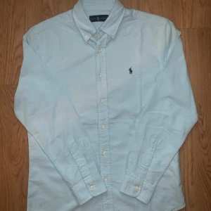 Ljusblå skjorta från Ralph Lauren - Snygg ljusblå skjorta från Ralph Lauren med klassisk button-down krage och broderad marinblå logga på bröstet. Skjortan har långa ärmar och vita knappar framtill. Perfekt för dig som gillar stilrena och tidlösa plagg.