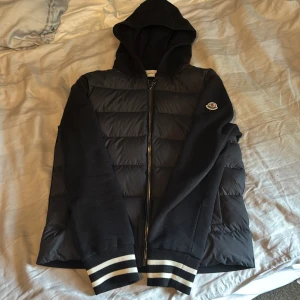 Moncler cardigan - Svart cardigan från Moncler med huva med ribbade muddar med vita ränder. Jackan har quiltad framsida, dragkedja och Moncler-logga på ärmen. Materialmix med polyester och bomull, perfekt för dig som gillar streetstyle och snygga detaljer. Nypris 7800 och skicket är som nytt.