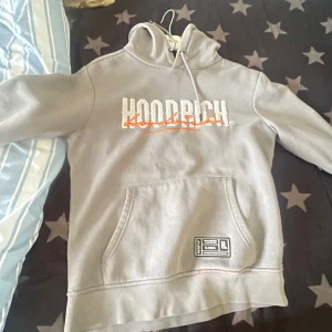 Grå hoodie från Hoodrich med tryck - Säljer en grå hoodie från Hoodrich i storlek S. Tröjan har stor vit och orange logga på både fram- och baksidan, samt en magficka och snörning i huvan. Materialet är mjukt och känns som bomull och polyester. Snygg streetwear-vibe med coola detaljer och patch vid fickan.