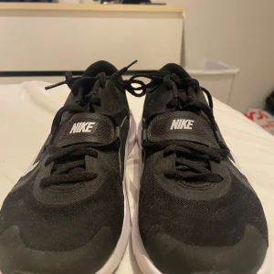 Svarta Nike sneakers med vit sula - Säljer ett par svarta Nike sneakers med vit sula och klassisk swoosh på sidan. Skorna har snörning och är tillverkade i syntetmaterial med mesh-överdel för bra ventilation. Perfekta för dig som gillar sportig stil och vill ha bekväma skor till vardagen.