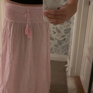 Ljusrosa långkjol med snörning - Supersöt ljusrosa långkjol med bred smock i midjan och dekorativ snörning med tofsar. Kjolen har ett diskret paisleymönster och är gjord i ett lätt, luftigt material som känns skönt mot huden. Perfekt för varma dagar och ger en chill vibe till din outfit. Använd få gånger. Kan ta ljusa bilder om de önskas. Storlek 158/164, lite kort i benen på mig som e 171 cm💕