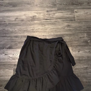 Svart omlottkjol med volang från ONLY - Svart omlottkjol från ONLY i storlek S. Kjolen har snygga volangdetaljer längs nederkanten och knytband i midjan för en justerbar passform. Perfekt för dig som gillar en enkel men trendig look.