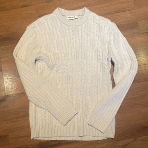 Ljusbeige stickad tröja från Kappahl - Säljer en ljusbeige stickad tröja från Kappahl i storlek S. Tröjan har ett snyggt flätmönster framtill och ribbade muddar vid hals, ärmslut och nederkant. Materialet är 70% polyester, 25% viskos och 5% ull. Perfekt för dig som gillar klassisk och enkel stil.