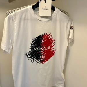 Vit Moncler t-shirt med  tryck. - Vit t-shirt från Moncler med ett stort  tryck i svart och rött på bröstet. Klassisk rund hals och korta ärmar. Moncler-logga broderad på vänster ärm. Mjuk bomullskvalitet som känns skön mot huden. Perfekt för dig som gillar stilrena designer med en twist. Köpt på NK. Ordinarie pris 2149kr.