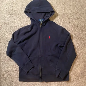 Mörkblå  zip-up hoodie från Polo Ralph Lauren - Snygg mörkblå hoodie från Polo Ralph Lauren med klassisk röd logobrodyr på bröstet. Tröjan har huva, dragkedja framtill och två fickor. Storlek L i barnstorlek (Om ni vill se kvitto fråga i privatmeddelande!) 