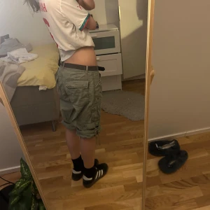 Gröna cargo shorts med snörning - Säljer ett par gröna cargo shorts med flera fickor och coola snörningsdetaljer vid bensluten. De har låg midja och är tillverkade i ett slitstarkt bomullsmaterial. Perfekt för dig som gillar streetstyle.
