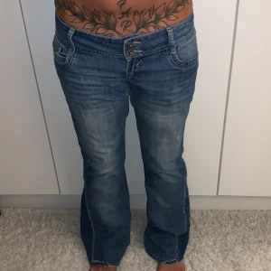Blå bootcut jeans från Dolce & Gabbana - Säljer ett par blå jeans  med bootcut passform och låg midja. Jeansen har slitningar, dubbla knappar i midjan och snygga detaljer på bakfickorna. Materialet är klassisk jeans med en skön känsla och cool tvätt. Det fanns ett hål som jag lagade, det syns på bilden.
