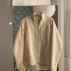 Beige teddyjacka från H&M Move - Mysig beige teddyjacka från H&M Move med hög krage och hel dragkedja framtill. Jackan har en bröstficka med dragkedja och är tillverkad i mjuk polyester. Perfekt för dig som gillar chill och stilren look.