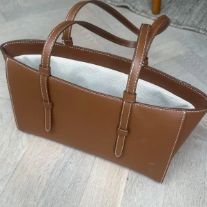 Brun handväska i ”skinn” med vita sömmar - Snygg brun handväska med stilrena vita sömmar och två rejäla handtag. Väskan har en avlång, rektangulär form. Insidan är ljus och kontrasterar snyggt mot det bruna yttermaterialet. Perfekt för dig som gillar klassisk och clean design.