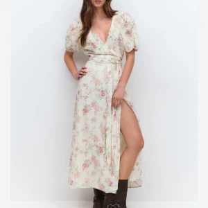 Blommig omlottklänning från Gina Tricot - Supersöt långklänning i ljus färg från Gina Tricot med blommigt mönster i rosa, grönt och beige. Klänningen har puffiga korta ärmar, v-ringning och omlott med knyt i midjan. Materialet är lätt och känns som bomull, perfekt för sommarens alla tillfällen.