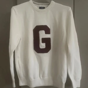 Vit stickad tröja med G från GANT - Vit stickad tröja från GANT med stor mörkröd G på bröstet. Rund hals med ribbad kant och diskret randig detalj. Långärmad och mjuk bomullskvalitet, perfekt för chill höst- och vinterdagar. Klassisk och clean look med college-vibe. Det är junior storlek men passar S