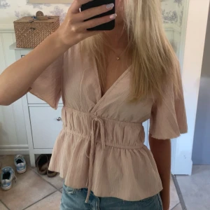 Beige omlottopp med knyt i midjan - Säljer denna supersöta beige/rosa omlottopp med korta ärmar och v-ringning. Säljer då den aldrig kommit till användning💕