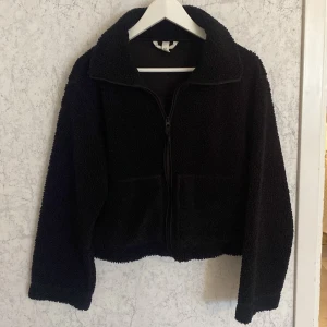 Svart teddyjacka med dragkedja - Mysig svart teddyjacka med hög krage och hel dragkedja framtill. Jackan har två stora fickor och är tillverkad i fluffigt fleece-material som ger en cool och avslappnad vibe. Perfekt för att hålla värmen under kyliga dagar i höst och passar till de flesta outfits. Från H&M storlek S.