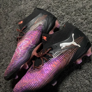Puma Future Z lila/svart fotbollsskor - Säljer ett par Puma Future Z fotbollsskor i en fet lila och svart färg med orange detaljer. Skorna har snörning, hög strumpliknande krage och coolt mönster på ovansidan. Sulan har lila och svarta dobbar för gräsplan. Perfekt för dig som vill sticka ut på planen.