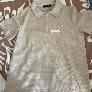 Prada polo beige pikétröja - Snygg beige pikétröja från Prada med diskret rutmönster och ikonisk metallbadge på bröstet. Tröjan har krage, korta ärmar och en stilren half zip framtill. Perfekt för dig som gillar clean och lyxig design. Priset kan diskuteras 🤩