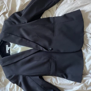 Svart kavaj från H&M, storlek 36 - Stilren svart kavaj från H&M i storlek 36. Klassisk modell med smal passform, enkel knappstängning och två fickor fram. Fodrad med vit, glansig insida. Perfekt för dig som gillar en clean och snygg look.