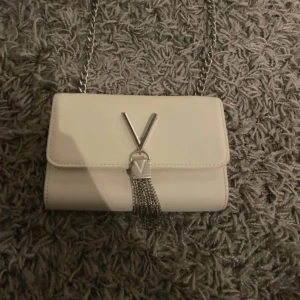 Vit axelväska från Valentino - Snygg vit axelväska från Valentino med silverkedja och dekorativt V-spänne framtill. Väskan har en stilren, rektangulär form och är gjord i skinn. Framsidan pryds av silverfärgade kedjefransar som ger en cool touch. Perfekt för dig som gillar exklusiva accessoarer.