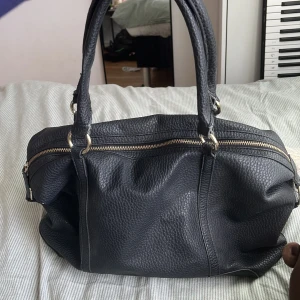 Svart handväska i skinn med gulddetaljer - Snygg svart handväska i kraftigt, strukturerat skinn med två rejäla handtag och guldfärgade ringar. Väldigt bra skick enda defekten är att de finns lite lösa trådar men de går enkelt att klippa bort. Väskan har en stor dragkedja upptill och flera fack, perfekt för att få plats med allt du behöver. Stilren och klassisk design som funkar till många outfits💖