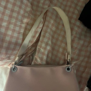 Rosa väska Lacoste - Så söt rosa lacoste väska med ljusrosa färg och vitt axelband, samt silvriga detaljer. Bra skick men syns att den är använd. Säljer direkt för 200kr.