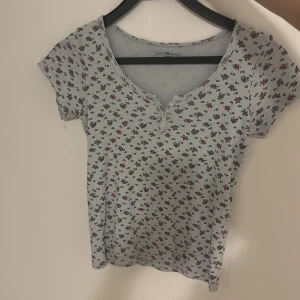 Blommig topp från Brandy Melville - Supersöt ljusblå topp från Brandy Melville med små blommor i rött och blått över hela. Toppen har korta ärmar, u-ringning med knappar framtill och är gjord i mjuk bomull.  Helt ny och oanvänd men utan prislapp🩷