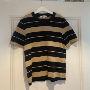 Randig t-shirt från COS i svart och beige - Snygg t-shirt från COS i storlek S med breda svarta och beige ränder. Klassisk rund hals och korta ärmar. T-shirten har en loose passform och är gjord i mjuk bomull som känns skön mot huden. Perfekt för dig som gillar stilrena och coola mönster.