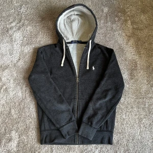 Hoodie från Ralph lauren - Säljer en snygg mörkgrå hoodie från Polo Ralph Lauren med dragkedja och justerbar huva. Tröjan har långa ärmar och en liten logga på bröstet. Perfekt för en avslappnad stil.