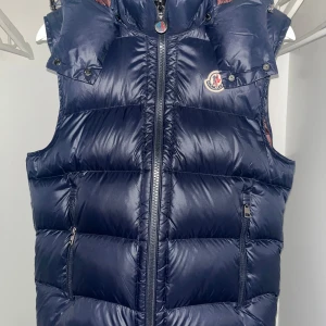 Moncler Bormes Väst - Snygg mörkblå dunväst från Moncler med dragkedja och huva. Västen har en quiltad design och är perfekt för vår, samt sommardagar. Insidan har ett unikt tryck med seriemotiv. Perfekt för dig som vill ha stil och funktion i ett. Storlek S motsvarar ”1” i moncler.                          (OBS!)         INGA BYTEN‼️‼️                                                                     Priset kan diskuteras om det sker snabbt och smidigt😇KVITTO FINNS! 
