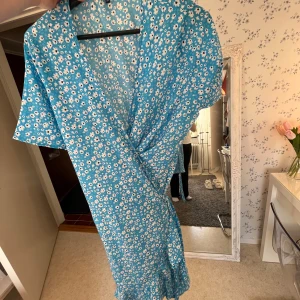 Blommig omlottklänning i blått - Säljer en söt blå omlottklänning med vita blommor. Klänningen har korta ärmar och volangdetaljer vid fållen. Perfekt för sommarens alla tillfällen!!