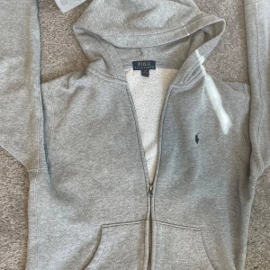 Grå hoodie från Ralph Lauren - Säljer en stilren grå hoodie från Ralph Lauren med dragkedja och huva. Tröjan har långa ärmar och en liten broderad logotyp på bröstet. Den är något nopprig men inget man märker av, det gör det snyggt! Perfekt för en avslappnad look. Storleken är L/G 14-16 160/80 160