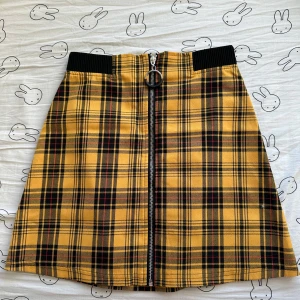 Gul rutig kjol med dragkedja från New Yorker - Snygg gul rutig kjol från New Yorker med dragkedja framtill. Kjolen har ett elastiskt svart band i midjan för extra komfort. Perfekt för att ge en färgglad touch till din outfit!