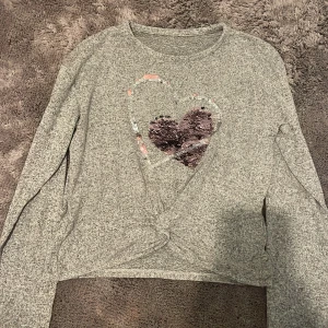 Grå tröja med paljetthjärta - Säljer en grå tröja med ett stort paljetthjärta på framsidan. Tröjan har långa ärmar och en knutdetalj framtill. Kommer från Lindex i storleken 146-152. Mycket fint skick endast använd ett fåtal gånger.