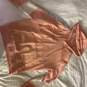 Rosa hoodie från Fila - Säljer en snygg rosa hoodie från Fila med vit logga på bröstet och Fila-band längs ärmarna. Tröjan har en stor ficka framtill och en bekväm huva med dragsko. Perfekt för en avslappnad stil!