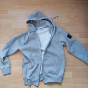 Grå hoodie från Stone Island - Säljer en grå hoodie från Stone Island med dragkedja och huva. Tröjan har en broderad logotyp på ärmen och är tillverkad i mjukt material. Perfekt för en avslappnad stil.