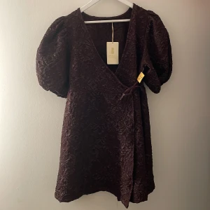 Mörklila omlottklänning från Pernilla Wahlgren - Snygg mörklila omlottklänning från Pernilla Wahlgren med puffiga kortärmar och ett vackert texturerat mönster. Klänningen har en v-ringad design och knytband i midjan för en smickrande passform. Perfekt för att sticka ut med stil!