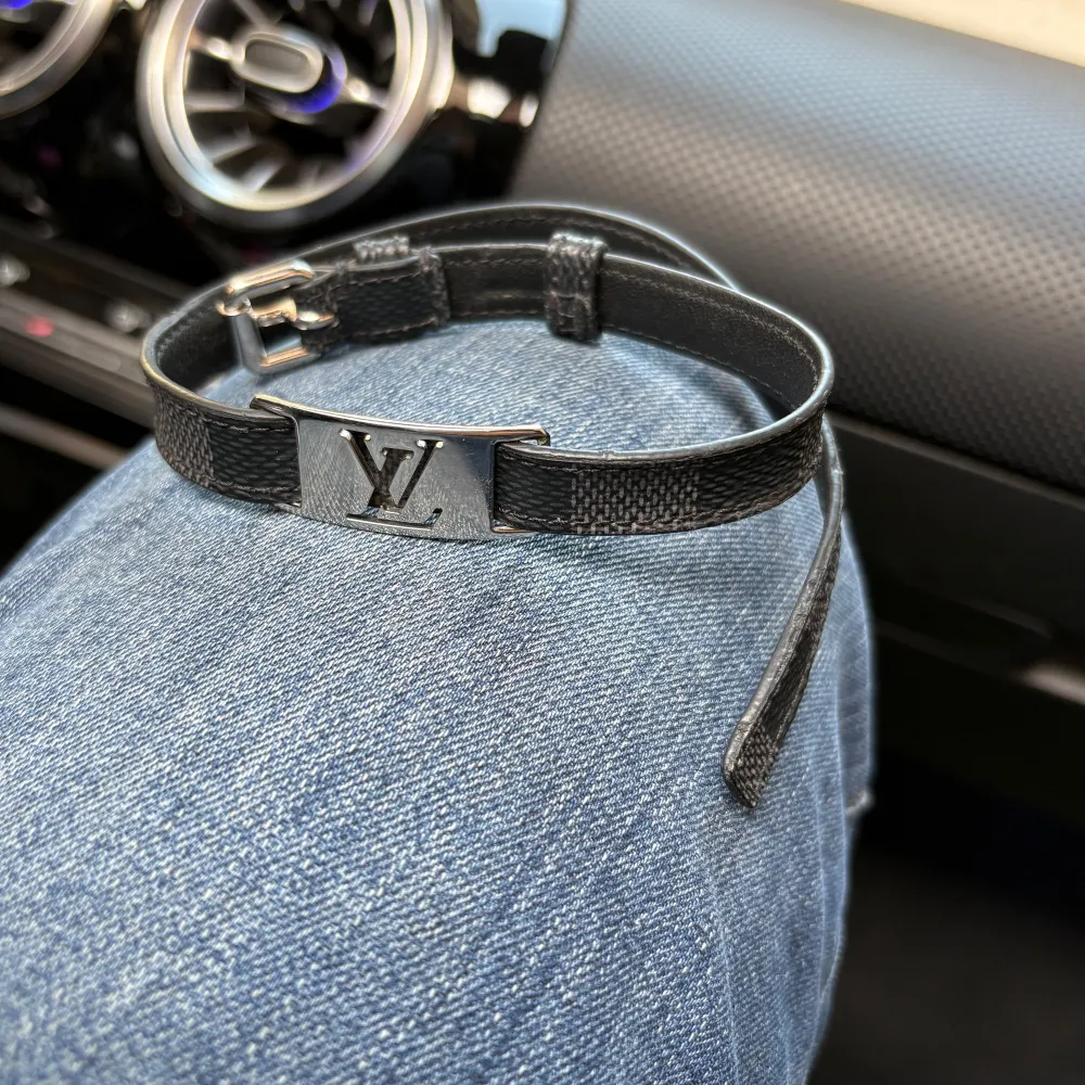 Snyggt svart armband från Louis Vuitton med en stilren silverdetalj i form av märkets logotyp. Armbandet har en justerbar rem och en elegant design som passar perfekt till både vardag och fest. Asusteet.