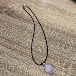 Halsband med lila sten - Snyggt halsband med en rå lila sten som hänge. Stenen är fäst på en tunn svart rem. Perfekt för att ge en naturlig touch till din stil.