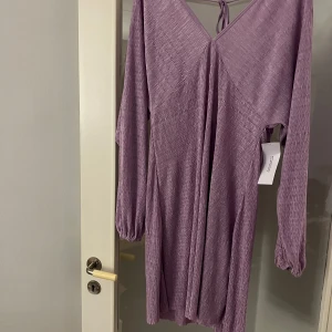 Klänning  - Snygg lavendel klänning från Bubbleroom med v-ringning och knytning i ryggen. Klänningen har långa ärmar och en mjuk, plisserad textur som ger en elegant look. Perfekt för att sticka ut med stil!