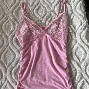 Rosa linne med spetsdetaljer - Säljer ett sött rosa linne med spetsdetaljer och smala axelband. Linnet har en liten rosett framtill och är perfekt för en varm dag. 