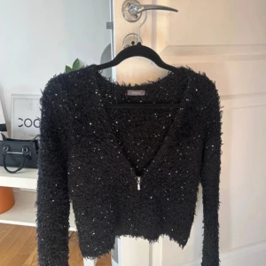 Svart glittrig kofta från Yessica - Snygg svart kofta från Yessica med glittriga detaljer och dragkedja framtill. Perfekt för att lägga till lite glamour till din outfit. Koftan har en v-ringad design och långa ärmar.