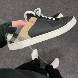Burrberry skor - Snygga svarta sneakers med ett klassiskt rutigt mönster på hälen. Skorna har vita snören och en vit sula som ger en stilren look. Perfekta för en avslappnad stil.