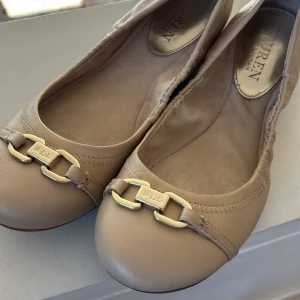 Beige ballerinaskor från Lauren Ralph Lauren - Snygga beige ballerinaskor från Lauren Ralph Lauren med guldfärgade detaljer framtill. Skorna har en rund tå och är tillverkade i skinn, vilket ger en elegant och stilren look. Perfekta för en chic och bekväm stil.