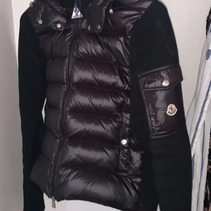 Svart dunväst från Moncler - Snygg svart dunväst från Moncler med stickade ärmar och dragkedja framtill. Västen har en quiltad design och en praktisk ficka på ärmen med knappdetalj. Perfekt för kyligare dagar. Kvitto finns och pris kan diskuteras vid snabbäffar 