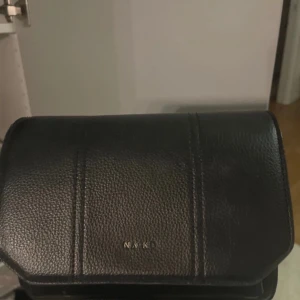 Svart axelväska från NA-KD - Snygg svart axelväska från NA-KD i skinnimitation. Väskan har en praktisk dragkedja och en stilren design som passar till många tillfällen. Perfekt för att bära dina nödvändigheter med stil.