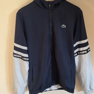 Blå vindjacka från Lacoste -  Den riktig bra för sommaren Snygg blå från Lacoste med dragkedja och huva. Jackan har vita och grå ärmar med Lacoste-logga på ärmarna. Perfekt för vår och höst. (VATTEN TÄTT)