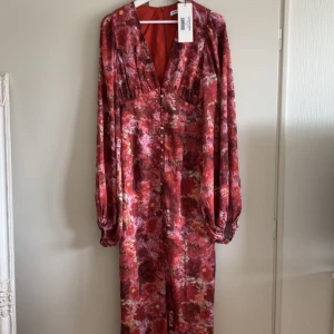 Positano  ADOORE - Säljer en vacker blommig långklänning från ADOORE i röd och rosa nyans. Klänningen har långa ärmar och en v-ringad design med knappar framtill. Aldrig använd med prislapp kvar.
