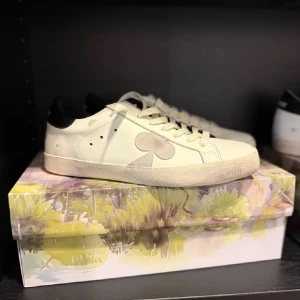 The gallerist skor - Stilrena vita sneakers med en svart detalj på hälen och ett unikt mönster på sidan. Skorna har snörning och en rund tå, vilket ger en klassisk look. Perfekta för en avslappnad stil.