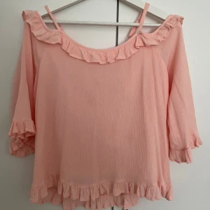 Rosa topp med volang från Lindex - Säljer en söt rosa topp från Lindex med volangdetaljer vid halsringning och ärmslut. 