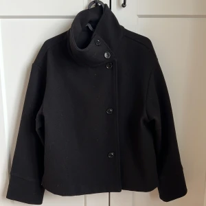 Svart kappa från Gina Tricot - Snygg svart kappa från Gina Tricot med hög krage och knappar framtill. Den har stora fickor på båda sidorna. 