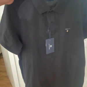 Pikétröjor från GANT - Säljer två stilrena pikétröjor från GANT. En i marinblått och en i vitt med svarta detaljer. Båda har korta ärmar och klassisk krage med knappar. Perfekta för en avslappnad men snygg look.